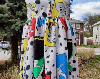 Puede incluir: Un vestido blanco sin mangas con cuello negro y ribete amarillo. El vestido tiene un diseño de patchwork colorido con perros dálmatas y varios colores, incluyendo rojo, azul, verde y amarillo.
