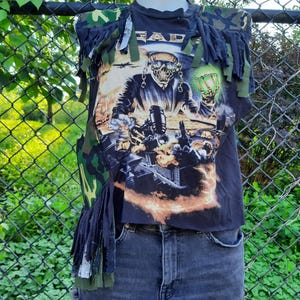 May include: A sleeveless black t-shirt with a graphic print of a skeleton wearing sunglasses and a suit, holding a glowing green object. The shirt features camouflage fabric accents on the shoulders and sides, with long fabric fringe detailing throughout.