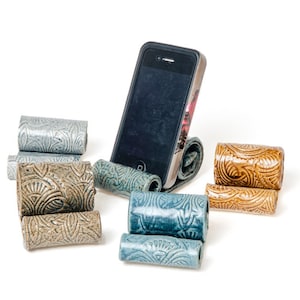May include: A black smartphone resting on a blue ceramic stand with a textured, embossed pattern. Surrounding the phone are several other ceramic stands in various colors including light blue, tan, and brown, all featuring similar embossed designs.