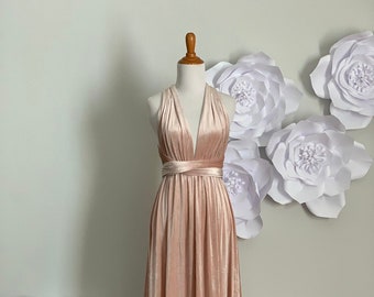 Könnte beinhalten: Ein langes, blushfarbenes Samtkleid mit einem Halterneck und einer Wickel-Taille. Das Kleid wird auf einer Schaufensterpuppe vor einem weißen Hintergrund mit Papierblumen präsentiert.