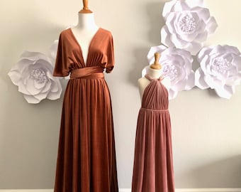 May include: Two velvet maxi dresses, one in a burnt orange color and one in a dusty rose color, on mannequins against a white wall with white paper flowers.