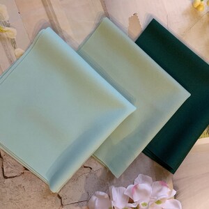 May include: Three fabric squares in different colors: light blue, green, and dark green. The squares are folded and stacked on top of each other.