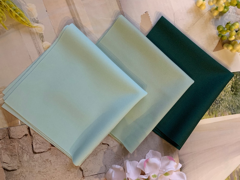 May include: Three fabric squares in different colors: light blue, green, and dark green. The squares are folded and stacked on top of each other.