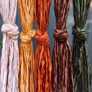 Pode incluir: Cinco lenços de seda em várias cores pendurados verticalmente, cada um amarrado no topo. As cores incluem rosa pálido, dourado, laranja, castanho e verde-oliva. O tecido tem uma aparência texturizada e plissada.