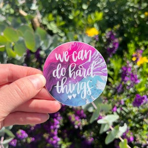 May include: A round sticker with the text "We can do hard things" in white script. The background features a watercolor design with pink, purple, and blue hues. Two small white hearts are at the bottom.