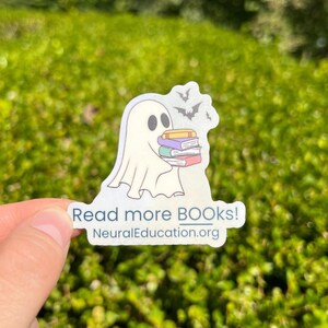 May include: A sticker featuring a cartoon ghost holding a stack of books. The ghost is white with black eyes and is carrying books with yellow, purple, and red covers. The sticker has the text "Read more BOOKS!" and "NeuralEducation.org".