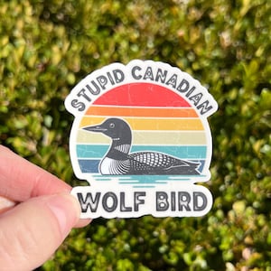 May include: A sticker featuring a loon illustration against a sunset backdrop with the text "STUPID CANADIAN" above and "WOLF BIRD" below. The sticker has a white border and is held against a green, leafy background.