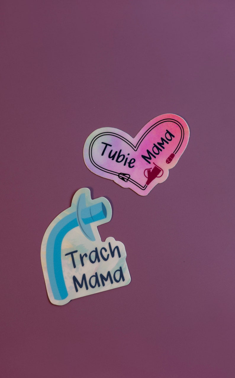 Może przedstawiać: Dwa naklejki z motywem medycznym. Pierwsza naklejka ma kształt serca z tęczowym gradientem i tekstem "Tubie Mama". Druga naklejka to niebiesko-biała ilustracja rurki tracheostomijnej z tekstem "Trach Mama".