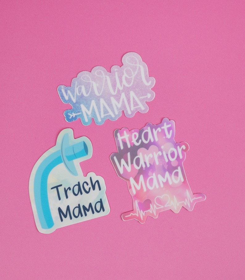 Może przedstawiać: Trzy kolorowe naklejki z tekstem "Warrior Mama", "Heart Warrior Mama" z sercem i linią EKG oraz "Trach Mama" z niebieską ilustracją rurki tracheostomijnej.