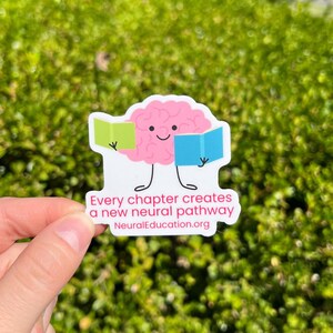 May include: A sticker featuring a pink brain character holding a green and blue book. The sticker reads "Every chapter creates a new neural pathway" and "NeuralEducation.org". The sticker is held in a hand against a green background.