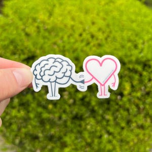 May include: A white sticker featuring a brain and a heart connected by a line. The brain is blue, and the heart is pink with a white interior. The sticker has a cartoonish design and is held against a green, blurred background.