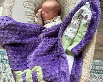 May include: A purple baby blanket with a green letter M on it. The blanket is soft and plush and has a textured surface. A baby is sleeping on the blanket.