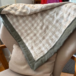 May include: A beige and white gingham throw blanket with a dark green border. The blanket is folded and draped over a chair.
