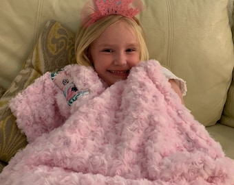 May include: A young girl wearing a pink "Happy Birthday" headband smiles while wrapped in a pink fluffy blanket.