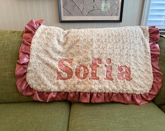 May include: A cream-colored, textured blanket with the name "Sofia" embroidered in a coral-pink color. The blanket has a ruffled, satin-like border in a matching coral-pink hue. The blanket is displayed on a green sofa.