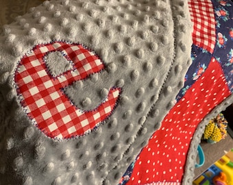 May include: A gray and red baby blanket with a red and white checkered letter E in the center. The blanket is made of soft, textured fabric.