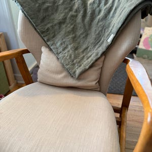 May include: A light green linen throw blanket draped over a beige armchair with a white pillow. The chair has wooden arms and legs.