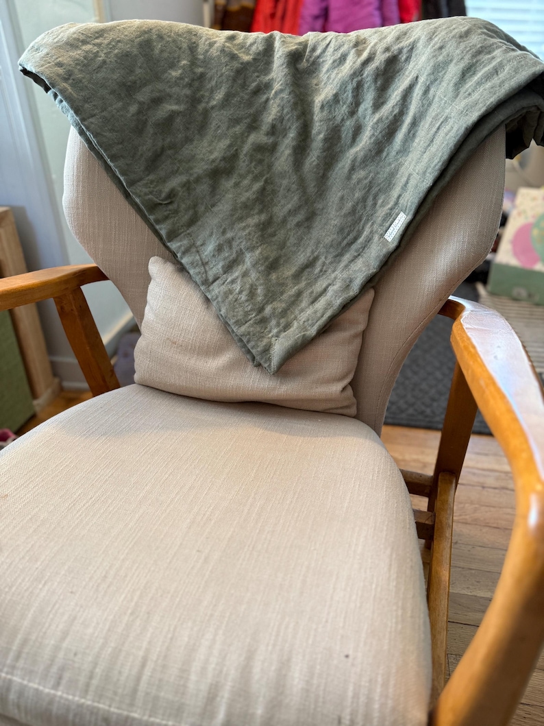 May include: A light green linen throw blanket draped over a beige armchair with a white pillow. The chair has wooden arms and legs.