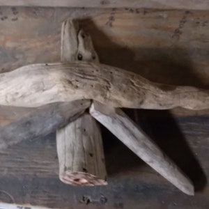 May include: A rustic, handcrafted driftwood sculpture. The artwork is assembled from several pieces of weathered wood, forming a cross-like shape with a central vertical piece and horizontal and angled arms. The wood has a natural, light brown color.