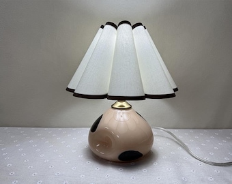 May include: A table lamp with a white fabric shade with a brown trim. The shade has a pleated design. The base of the lamp is a pink ceramic sphere with black polka dots.