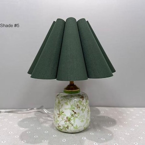 May include: A decorative table lamp with a green fabric shade and a floral patterned base. The shade is a deep green color and has a unique, pleated design. The base is white with a floral pattern and a gold-colored accent.