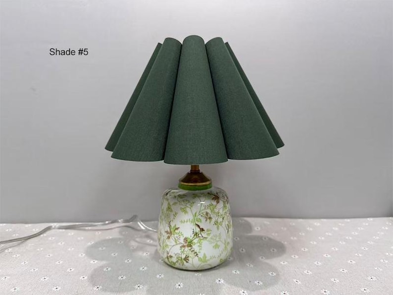 May include: A decorative table lamp with a green fabric shade and a floral patterned base. The shade is a deep green color and has a unique, pleated design. The base is white with a floral pattern and a gold-colored accent.
