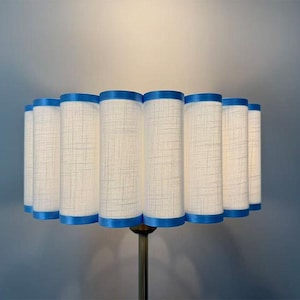 Peut inclure: Abat-jour cylindrique avec un tissu crème et une bordure bleue. L'abat-jour a un design plissé et est monté sur un pied en métal couleur laiton. La lampe est un luminaire décoratif.