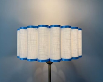 May include: A cylindrical lampshade with a cream-colored fabric and blue trim. The shade has a pleated design and is mounted on a brass-colored metal stand. The lamp is a decorative lighting fixture.