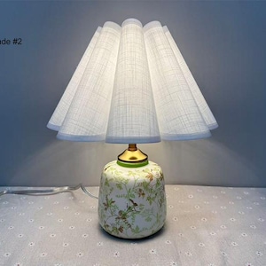 May include: A decorative table lamp with a white pleated shade and a floral patterned base. The base is a light green color with gold accents. The lamp is plugged in and has a white cord. The text "Shade #2" is visible.