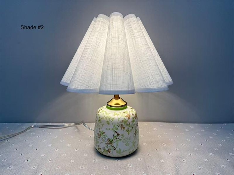 May include: A decorative table lamp with a white pleated shade and a floral patterned base. The base is a light green color with gold accents. The lamp is plugged in and has a white cord. The text "Shade #2" is visible.
