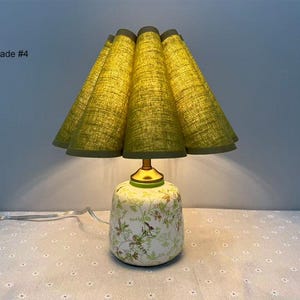 May include: A decorative table lamp with a green, pleated lampshade and a floral patterned base. The shade is a light green color, and the base is white with green and brown floral designs. The lamp has a gold-colored neck and a white power cord.