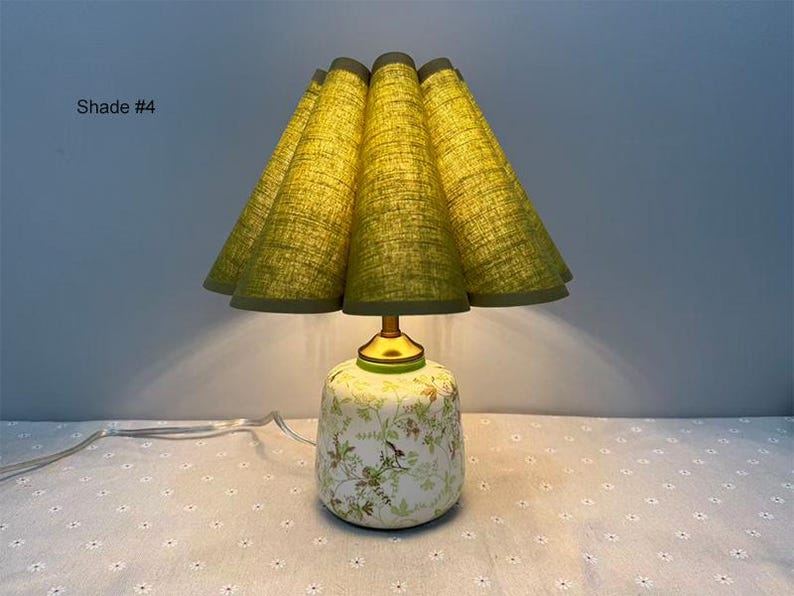 May include: A decorative table lamp with a green, pleated lampshade and a floral patterned base. The shade is a light green color, and the base is white with green and brown floral designs. The lamp has a gold-colored neck and a white power cord.