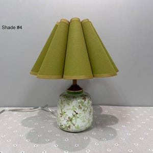 May include: A decorative table lamp with a green lampshade and a floral patterned base. The lampshade is a light green color with a gold trim. The base is white with green and brown floral designs. The text "Shade #4" is in the upper left corner.