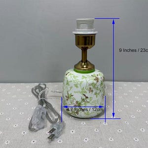 May include: A decorative ceramic lamp with a floral pattern in green and brown. The lamp is 9 inches tall and 4.7 inches wide. It has a brass-colored neck and a white lamp socket. A power cord with a plug is attached.
