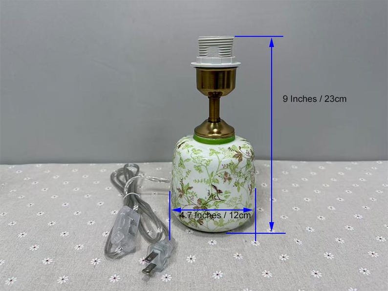 May include: A decorative ceramic lamp with a floral pattern in green and brown. The lamp is 9 inches tall and 4.7 inches wide. It has a brass-colored neck and a white lamp socket. A power cord with a plug is attached.