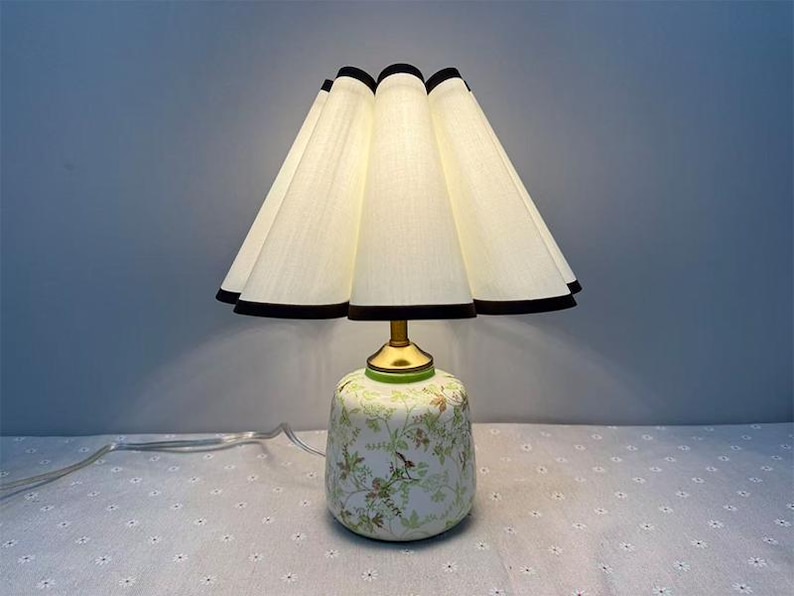 May include: A decorative table lamp with a white lampshade trimmed with black, and a floral patterned base. The lamp has a brass-colored neck and a white cord.