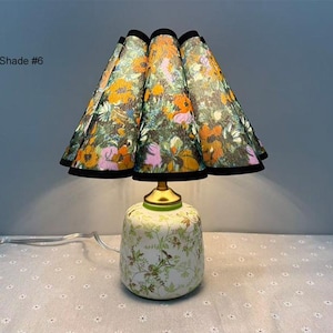 May include: A decorative table lamp with a floral patterned lampshade and base. The lampshade features a colorful floral design with orange, pink, and green flowers on a dark background. The base is white with a green floral pattern. The text "Shade #6" is visible.