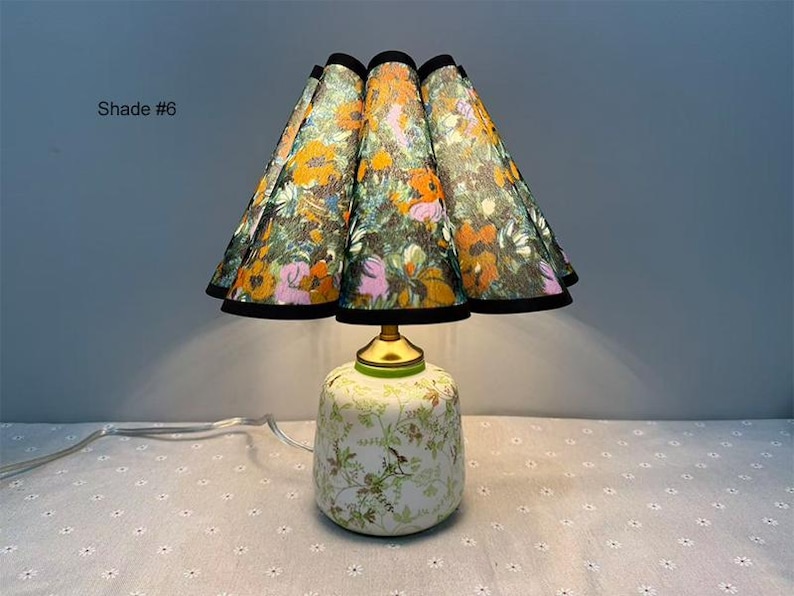 May include: A decorative table lamp with a floral patterned lampshade and base. The lampshade features a colorful floral design with orange, pink, and green flowers on a dark background. The base is white with a green floral pattern. The text "Shade #6" is visible.