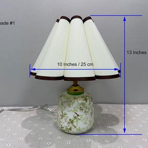 May include: A decorative table lamp with a white pleated shade, 10 inches (25 cm) wide, and a height of 13 inches (33 cm). The shade has a brown trim. The base is white with a floral design.