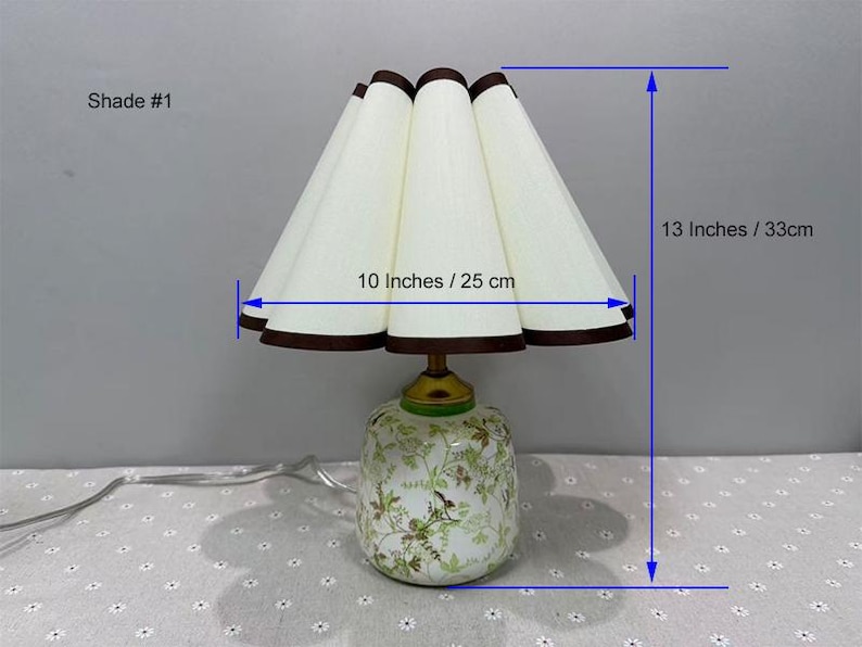 May include: A decorative table lamp with a white pleated shade, 10 inches (25 cm) wide, and a height of 13 inches (33 cm). The shade has a brown trim. The base is white with a floral design.