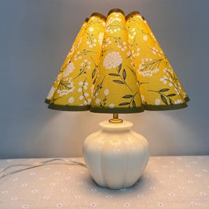 May include: A white ceramic table lamp with a pleated yellow lampshade featuring a floral pattern. The lampshade has a green trim.