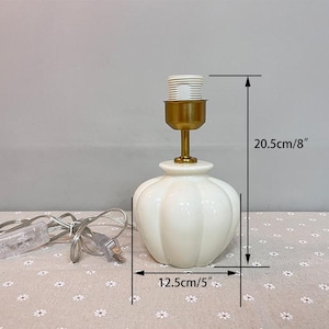 May include: A white ceramic table lamp with a gold-colored neck and bulb socket. The lamp base is 5 inches wide and 8 inches tall. The lamp has a power cord and is on a patterned surface.