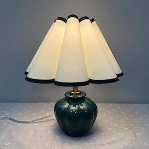 May include: A table lamp featuring a dark green ceramic base and a white lampshade with a black trim. The lampshade has a pleated design. The lamp is on a patterned surface.