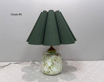 May include: A decorative table lamp with a green fabric shade and a floral patterned base. The shade is a deep green color and has a pleated design. The base is white with a floral pattern in green and brown. The text "Shade #5" is visible.