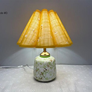 May include: A vintage-style table lamp with a yellow pleated shade and a floral patterned base. The shade is a bright yellow with a grid pattern and small white floral accents. The base is white with green and brown floral designs.