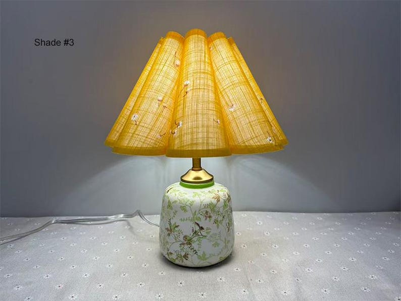 May include: A vintage-style table lamp with a yellow pleated shade and a floral patterned base. The shade is a bright yellow with a grid pattern and small white floral accents. The base is white with green and brown floral designs.