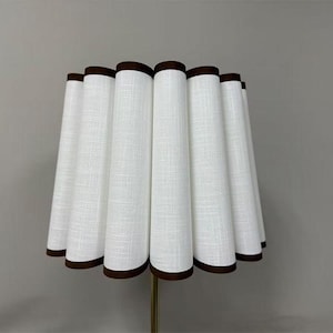 May include: A white lampshade with a pleated design and a dark brown trim. The lampshade has a cylindrical shape and is set against a neutral grey background. The lampshade is a decorative lighting element.