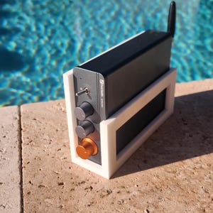 May include: A black electronic device with a silver antenna and control knobs, mounted in a white frame. The device has an orange knob and is set against a backdrop of a pool.