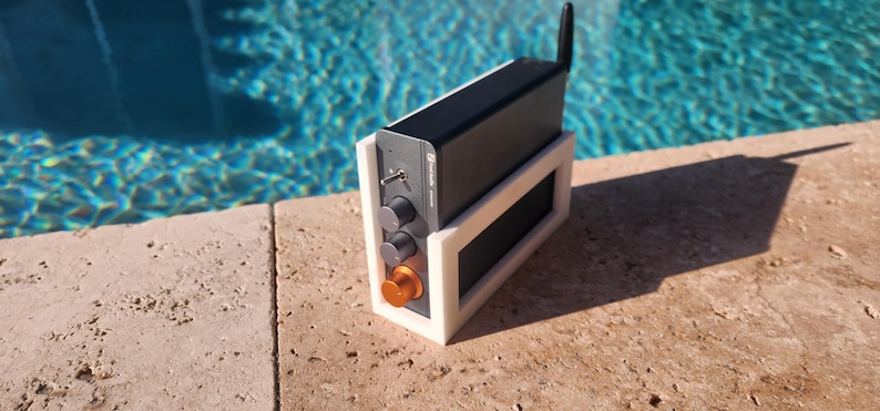 May include: A black electronic device with a silver antenna and control knobs, mounted in a white frame. The device has an orange knob and is set against a backdrop of a pool.