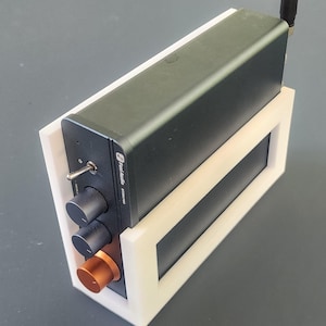 May include: A black audio amplifier with a silver toggle switch, two black knobs, and a copper-colored knob. The amplifier is mounted in a white frame and has a small antenna. The text "Fosi Audio BT30A PRO" is visible on the device.